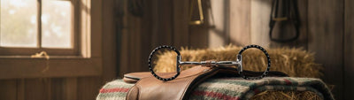 Myler 03 Black Dee Bit, a 5-inch sweet iron comfort snaffle, displayed on a brown leather saddle with a plaid blanket, atop a hay bale in a rustic barn.