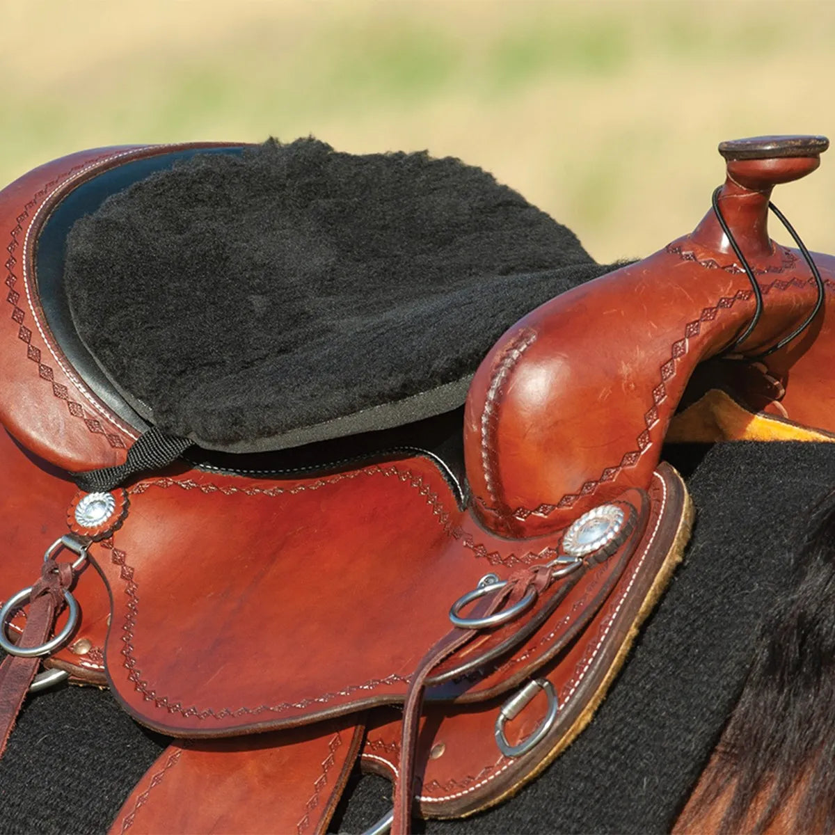 Black Cashel Fleece Tush Cushion seat pad installed on a chestnut leather western saddle with decorative tooling. This plush, non-slip fleece cushion offers superior comfort for trail riding and long hours in the saddle, featuring durable elastic straps that secure easily to the cantle and horn. Ideal equestrian accessory for western riders seeking a soft, cushioned seat.