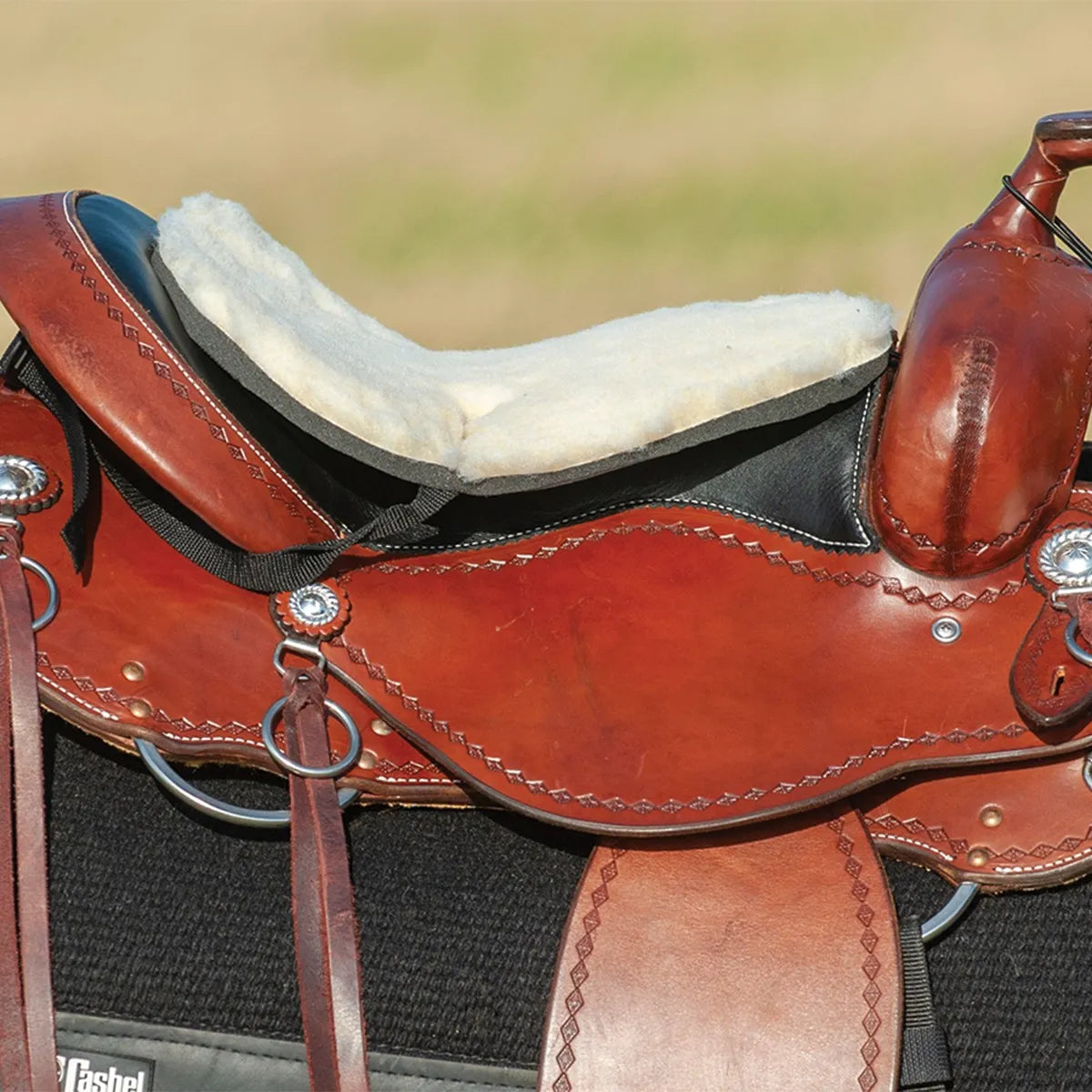 Cashel Fleece Tush Cushion in white fleece, designed for maximum comfort on long trail rides. This thick, padded seat cushion is shown securely attached to a brown leather western saddle with decorative tooling. Featuring a soft fleece top and black foam base, it contours perfectly to the saddle seat to provide riders with extra cushioning and pressure relief. Ideal western horse tack accessory for endurance and trail riding.