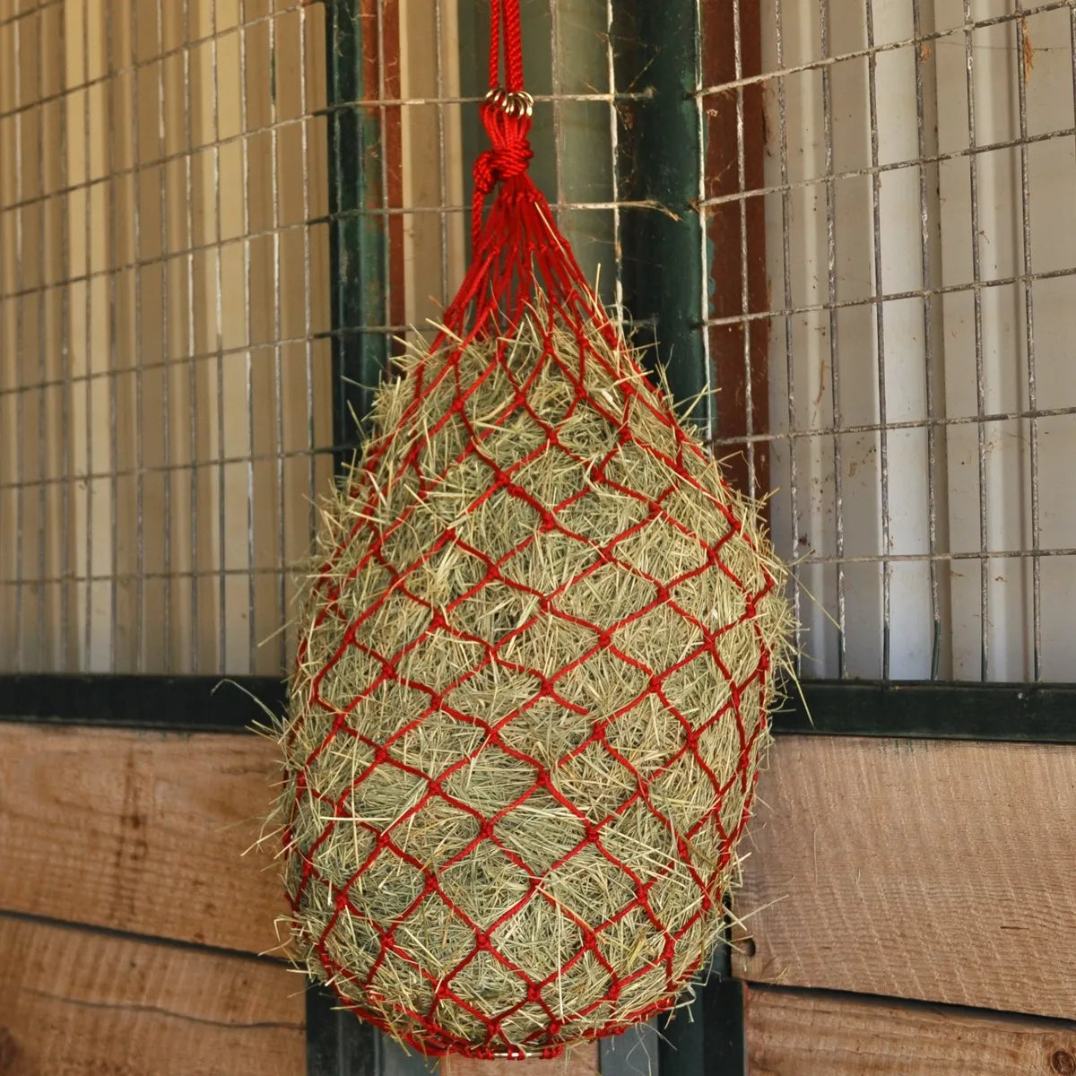 Red Cashel Hay Net slow feeder for horses filled with hay and hanging in a wooden barn stall. The small diamond mesh design helps reduce hay waste and promote slower eating for better equine health. This durable red hay bag features sturdy rope construction and metal rings for easy hanging, making it an essential piece of horse stable equipment for stalls or trailers.