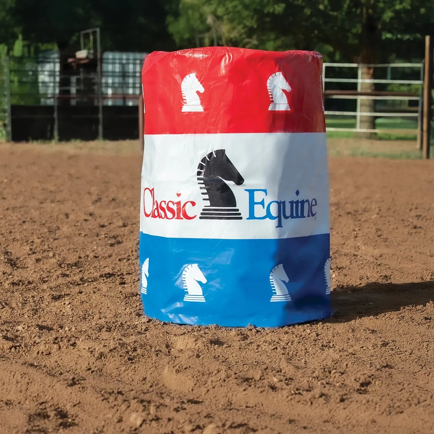 Classic Equine Barrel Cover featuring a patriotic red, white, and blue three-stripe design with the brand’s signature horse head logo. This durable equestrian training accessory is shown positioned in a dirt riding arena, ideal for barrel racing practice and professional competitions. Perfect for horse trainers and barrel racers looking for high-visibility, professional-grade barrel wraps to enhance their western arena setup.