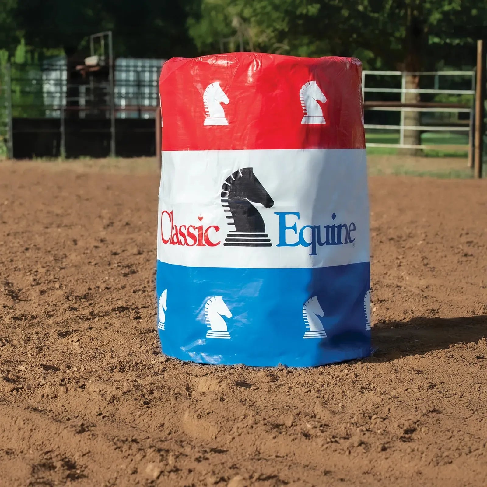 Classic Equine Barrel Cover featuring a patriotic red, white, and blue three-stripe design with the brand’s signature horse head logo. This durable equestrian training accessory is shown positioned in a dirt riding arena, ideal for barrel racing practice and professional competitions. Perfect for horse trainers and barrel racers looking for high-visibility, professional-grade barrel wraps to enhance their western arena setup.