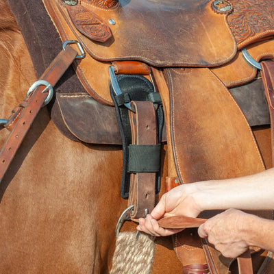 Classic Equine Cinch & Latigo Protector in black, shown installed on a brown leather Western saddle. This anti-rub and anti-itch padded accessory sits behind the latigo strap and cinch buckle to prevent skin irritation and galling. The close-up view shows a hand tightening the leather latigo, illustrating how the protector provides a comfortable barrier between the horse and the hardware. Ideal for sensitive horses and long trail rides.