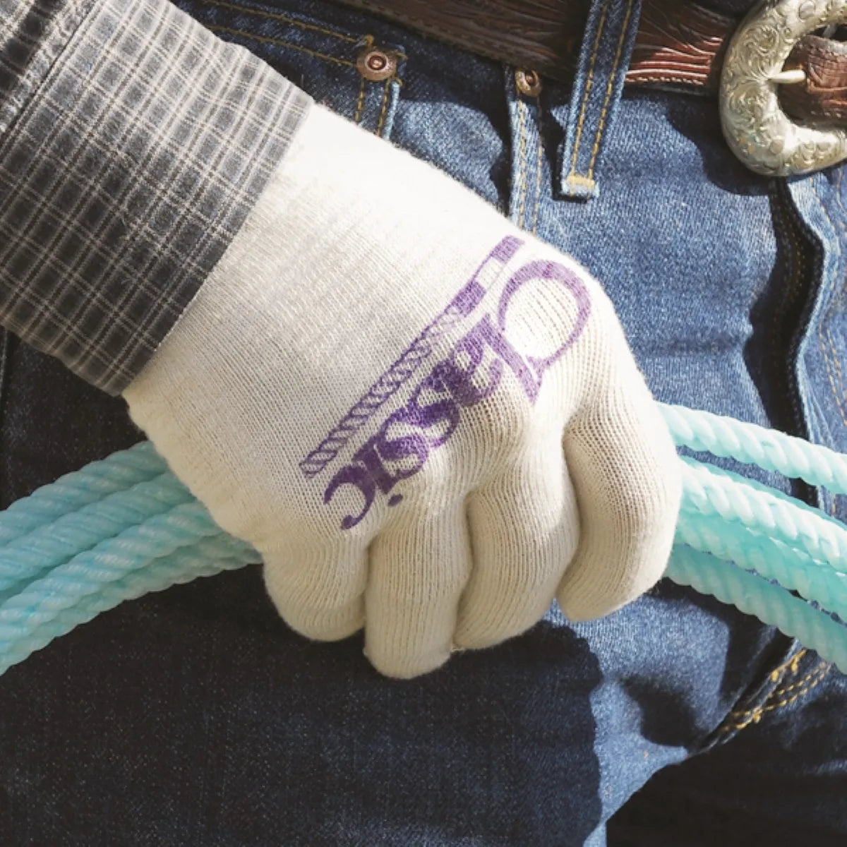 Classic Equine white knit roping glove featuring a purple brand logo, gripped around a professional turquoise rope. These high-performance rodeo gloves offer superior feel and durability for team roping. Shown in a Western setting against blue denim jeans and a silver belt buckle, they are essential equestrian gear for ropers seeking a secure grip and maximum protection.