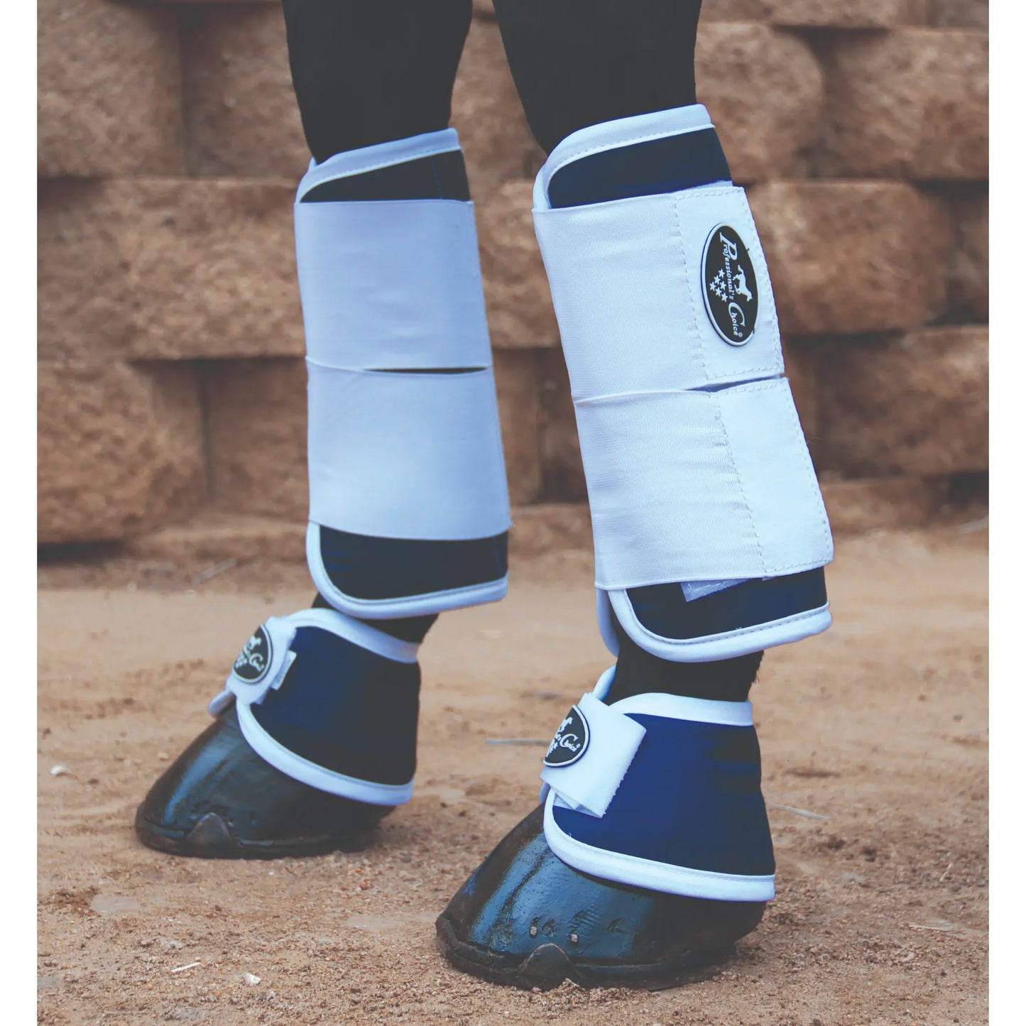 Professional's Choice Magnetic Tendon Boots and matching navy bell boots on a horse's front legs. These white and navy blue therapeutic boots offer advanced pain relief and injury prevention for equine athletes. Featuring a secure two-strap design for a perfect fit during recovery or protection, these boots are essential equestrian gear for horse therapy and daily leg protection, shown in a sandy arena with a rustic stone backdrop.