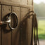 Myler 03 Black Dee Bit, a 5-inch sweet iron comfort snaffle, hangs on a dark wooden stable door with a blurred horse grazing in a sunlit green pasture in the background.