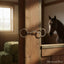Myler MB 06 Loose Ring Snaffle horse bit made of shiny silver metal, hanging on a weathered wooden barn post. Sunlight streams into the barn, with a blurred horse and hay bales in the soft-focus background, depicting an equestrian setting.