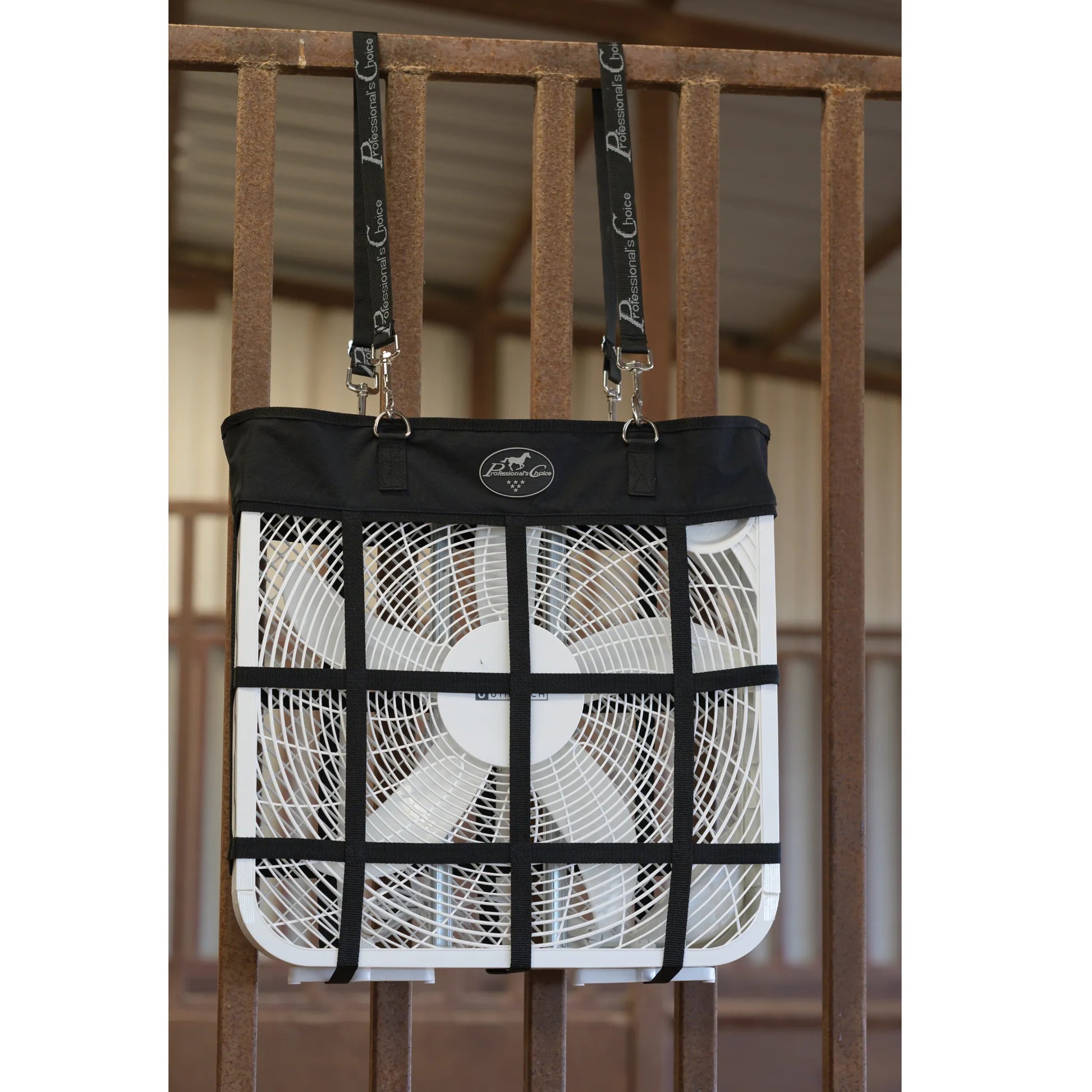 Professional's Choice Adjustable Fan Hanger in black, securely holding a standard white box fan against a metal stall gate. This heavy-duty equestrian fan holder features durable webbing in a grid pattern and adjustable straps with metal clips for easy mounting in horse stalls or barns. The top panel displays the Professional's Choice logo. Ideal for keeping horses cool and comfortable during hot summer days at the stable or horse shows.