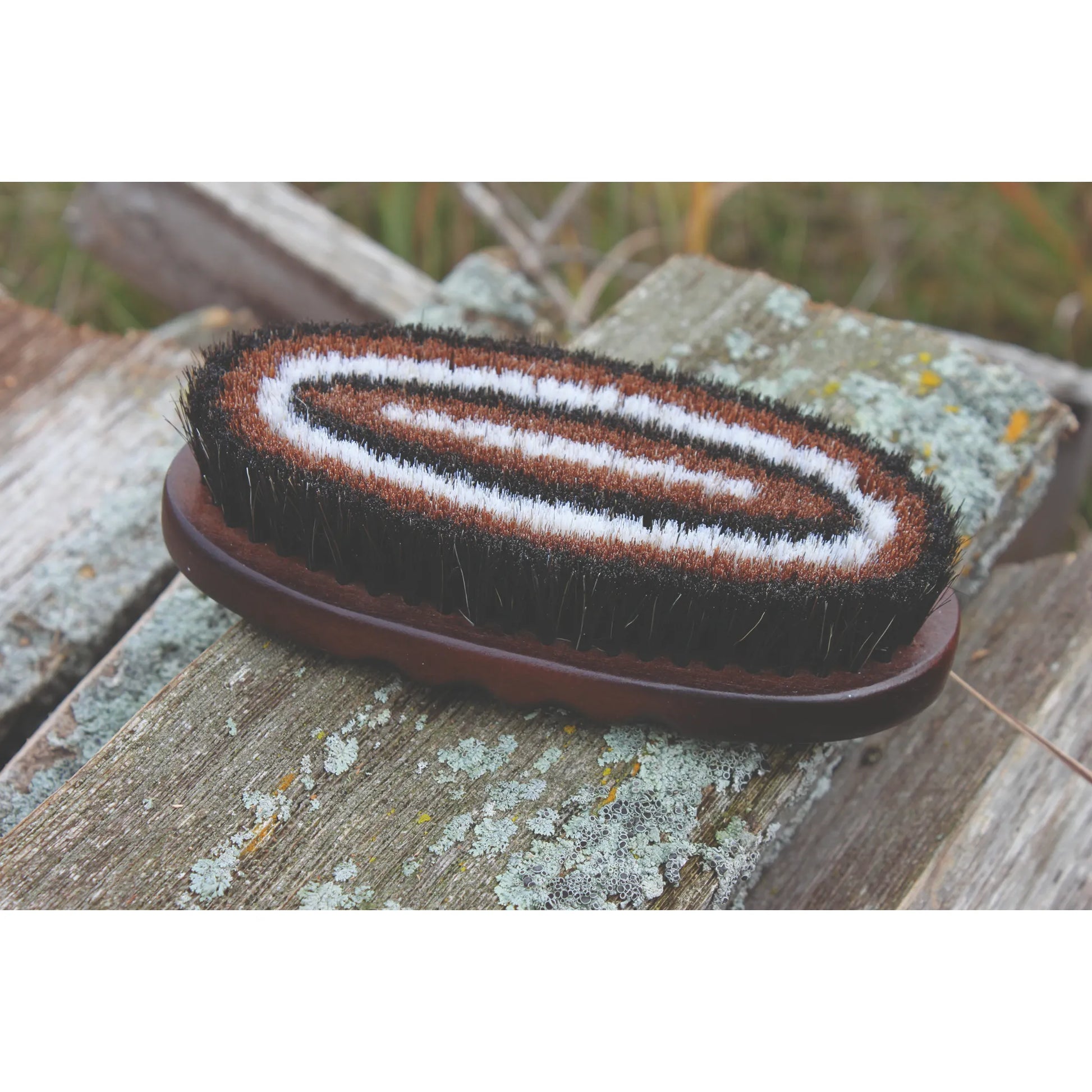 Professionals Choice Oval Wooden Horse Hair Brush for gentle equestrian grooming. Features a dark wood base with soft, dense bristles arranged in a stylish concentric pattern of black, brown, and white. This premium horse brush is ideal for finishing coats and daily care. Shown in a rustic outdoor setting, perfect for horse owners seeking high-quality, professional-grade grooming supplies.