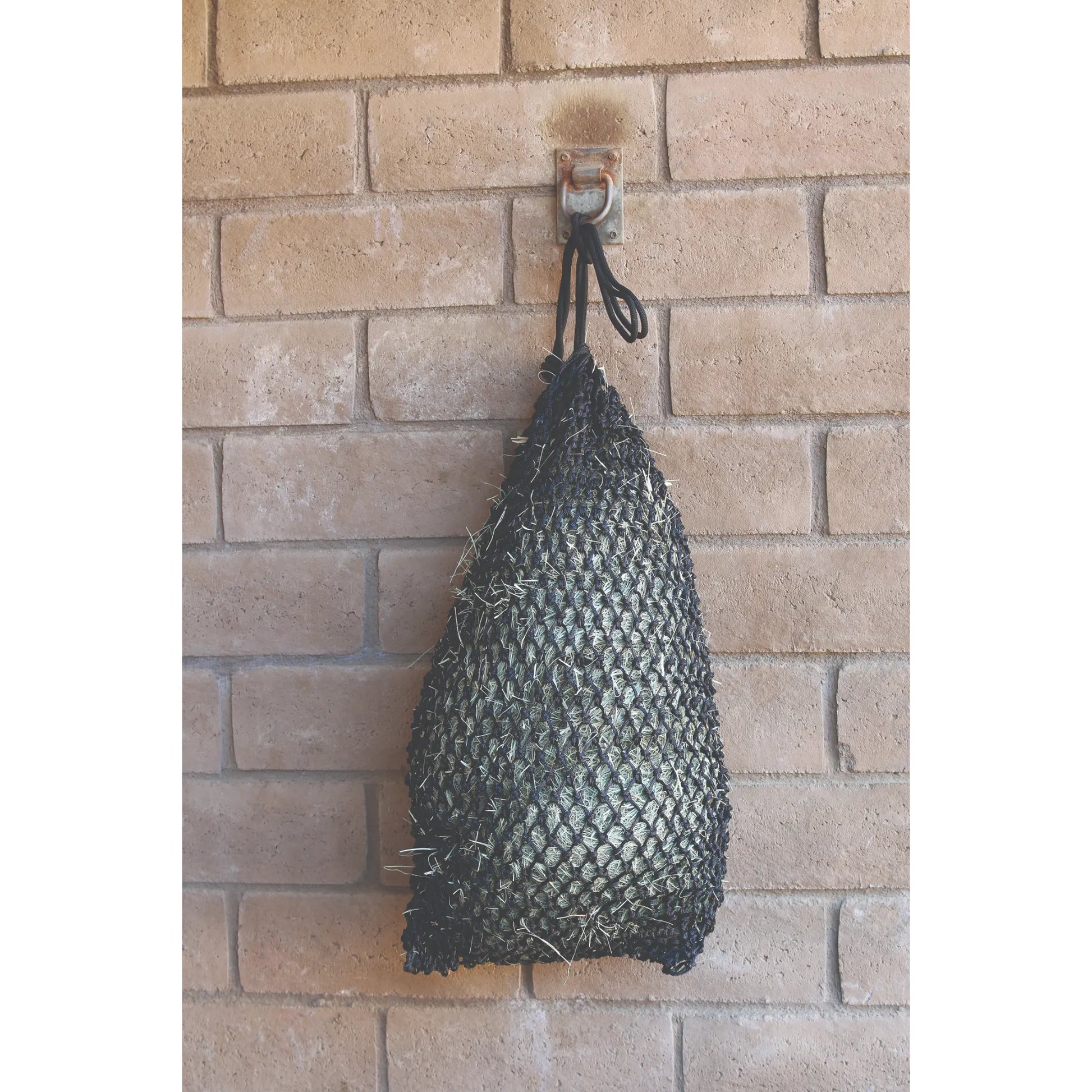 Professional’s Choice Slow Feed Hay Net in black, filled with hay and hanging against a brick wall. This durable, small-mesh horse feeder reduces waste and promotes healthy, slow grazing for better digestion. Ideal for stable or trailer use, this high-quality nylon hay bag keeps feed clean and off the ground, making it a must-have for equestrian care and efficient stable management.