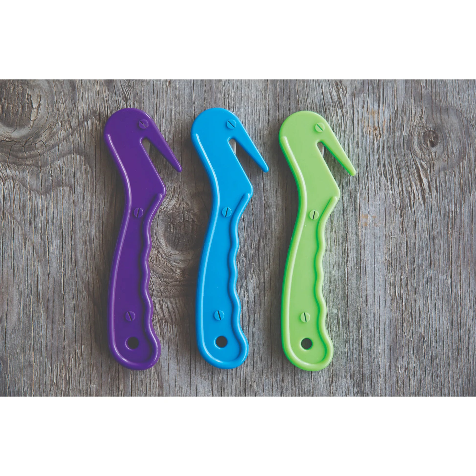 Professionals Choice Soft Touch Bale Cutters in vibrant purple, blue, and lime green. These ergonomic hay bale openers feature a recessed safety blade for protection and a soft-grip handle for comfort. A must-have barn tool for easily cutting twine and plastic wrap on hay bales, shown on a rustic gray wood background. Durable equestrian stable supplies perfect for daily farm chores and feed room organization.