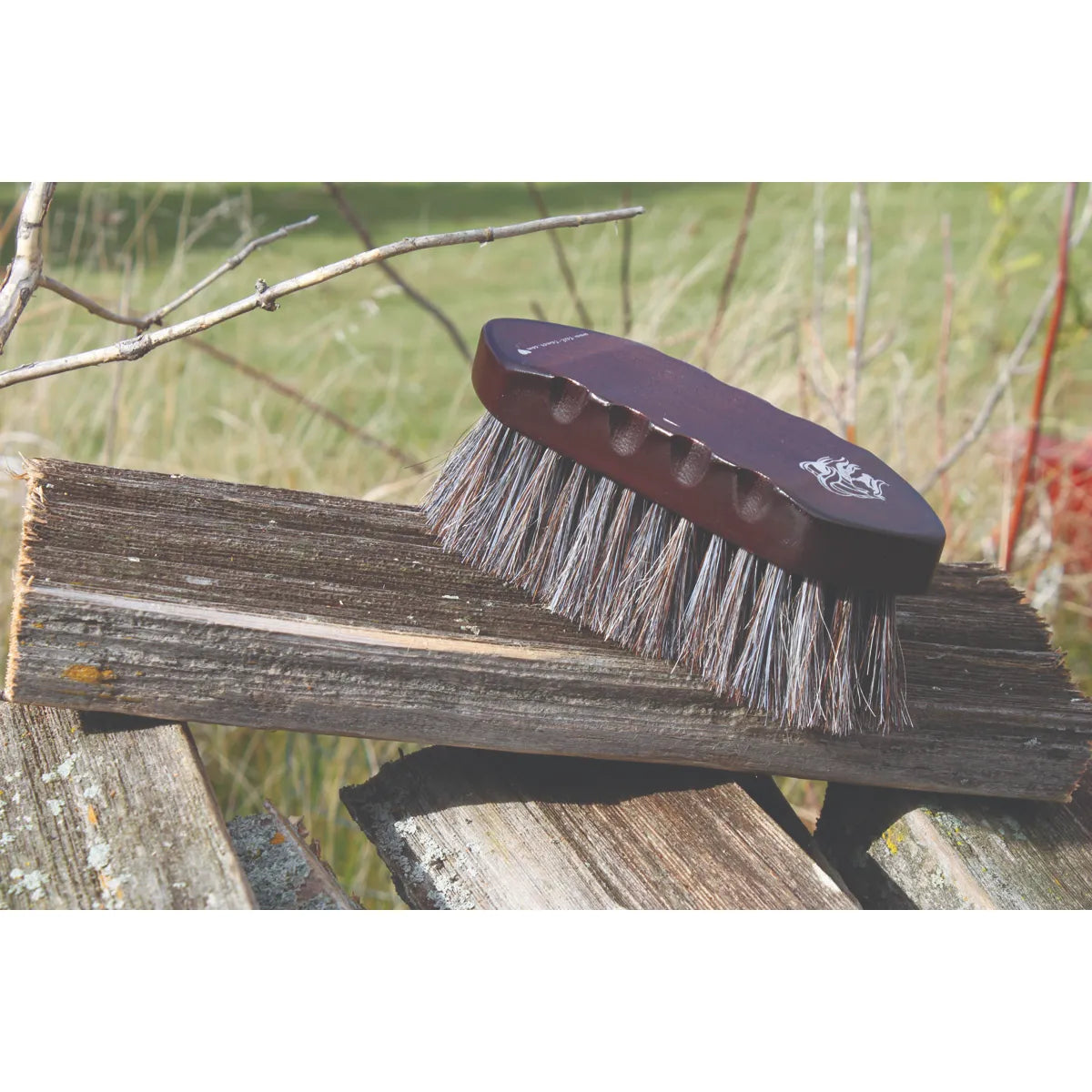 Professionals Choice Wood Series Small Horsehair Brush with a dark mahogany wood handle and ergonomic finger grooves. This professional horse grooming tool features dense, natural horsehair bristles for a high-quality finish. Shown resting on a rustic wooden plank outdoors, this durable and stylish brush is an essential addition to any equestrian grooming kit. Perfect for horse care enthusiasts looking for quality and elegance.