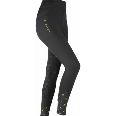 Shires Aubrion Porter Maids Winter Riding Tights in black, featuring a side phone pocket with gold branding and silicone grip on the inner legs for equestrian stability. These fleece-lined riding leggings include stylish gold-patterned ankle cuffs and a high-waist design, perfect for keeping young riders warm in the saddle during winter.